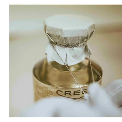 A close-up shows the top of a gold bottle with a textured silver cap tied with a silver cord, with the text CREED 1760 visible on the bottle.