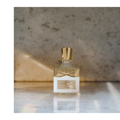 A clear glass bottle of Creed Aventus Cologne perfume with a silver cap rests on a marble surface, featuring the logo CREED and label text AVENTUS COLOGNE.