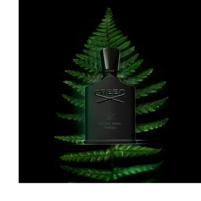A black perfume bottle with the text CREED GREEN IRISH TWEED sits in front of a large green fern leaf.