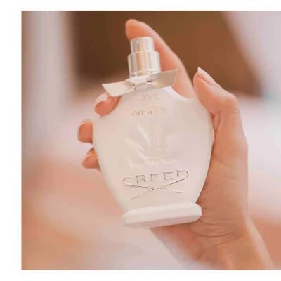 A hand holds a white perfume bottle with a light grey bow ribbon and the text LOVE IN WHITE CREED embossed on it.