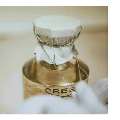 Close-up of a golden bottle, likely perfume, with a cloth-covered cap tied with a string, showing part of the bottles label with text CREED 1760.