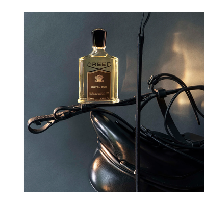 A Creed Royal Oud perfume bottle is displayed on black leather equestrian gear including a saddle and bridle, with visible text CREED ROYAL OUD DE PERE EN FILS DEPUIS 1760 FROM FATHER TO SON SINCE 1760 100ML 3.33 FL. OZ.