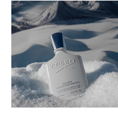 A white perfume bottle with a dark blue cap is nestled in glistening white snow, with blurry snow-covered mountains in the background, and the bottle displays text including CREED, SILVER MOUNTAIN WATER, DE PERE EN FILS DEPUIS 1760, FROM FATHER TO SON SINCE 1760, 100 ML, 3.33 FL. OZ.