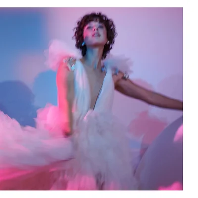 A person with dark curly hair and a white, deep V-neck ruffled dress with sparkling shoulder adornments looks up, captured with motion blur against a background of blue, pink, and purple light