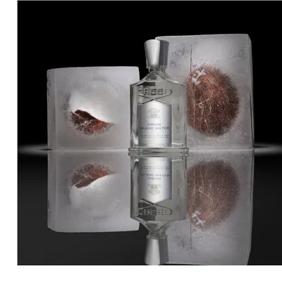 A Creed perfume bottle is shown standing between two ice blocks with pieces of coconut frozen inside, reflected on a dark surface below, with the label reading CREED VIRGIN ISLAND WATER DE PÈRE EN FILS DEPUIS 1760 FROM FATHER TO SON SINCE 1760 100ML 3.33 FL.OZ.