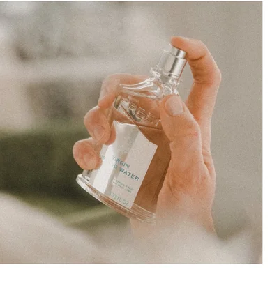 A hand holds a glass bottle of Creed Virgin Island Water perfume showing the label with text VIRGIN ISLAND WATER, FRANCAIS DEPUIS 1760 ENGLISH SINCE 1760, and 3.33 FL. OZ.