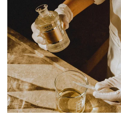 A person in white gloves pours liquid from a glass bottle labeled CREED ESIME IMPERIAL into a beaker.