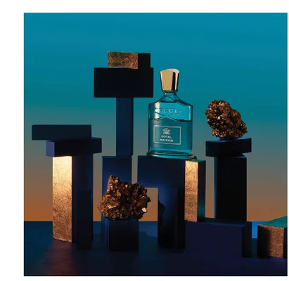 A blue Creed perfume bottle with a gold cap is displayed among dark blocks and golden rocks, with the text CREED ROYAL MAYFAIR visible on the label.