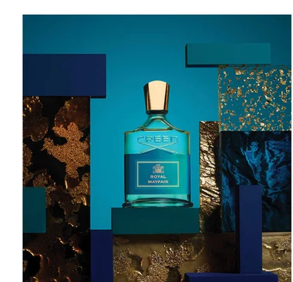 A Creed perfume bottle is centered on a dark green block surrounded by blue and gold abstract shapes, with text CREED ROYAL MAYFAIR on the bottle.