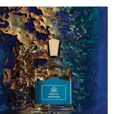 A Creed perfume bottle with a blue label featuring a crest symbol and the text ROYAL MAYFAIR is displayed against a textured gold and blue background, with CREED embossed on the bottle.