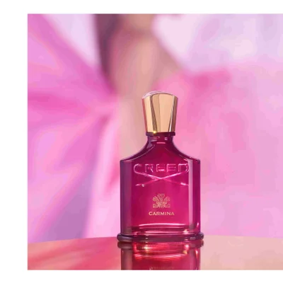 A pink perfume bottle with a gold cap sits on a reflective surface against a soft pink background with the text CREED CARMINA visible on the bottle.