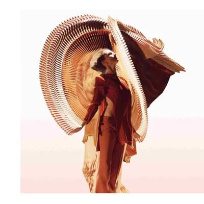 A woman in a red-orange outfit stands in front of a large layered circular structure.