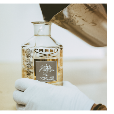 Close up of liquid being poured from a glass bottle labeled CREED into a glass beaker with a pipette in the background.