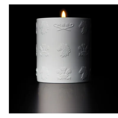 A lit white candle with embossed designs and the text CREED sits on a reflective surface in a dark setting