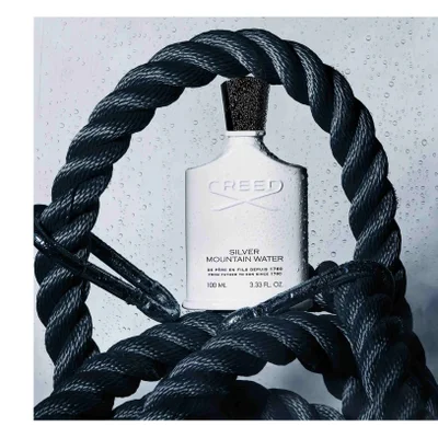 A white Creed Silver Mountain Water fragrance bottle with a black cap is covered in water droplets and surrounded by black rope, displaying the text Creed Silver Mountain Water De Père en Fils Depuis 1760 From Father to Son Since 1760 100 ML. 3.33 FL. OZ.