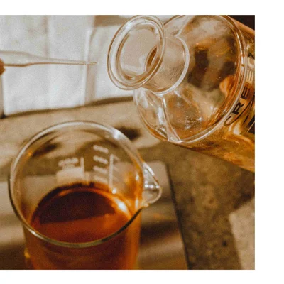 Close up of liquid being poured from a glass bottle labeled CREED into a glass beaker with a pipette in the background.