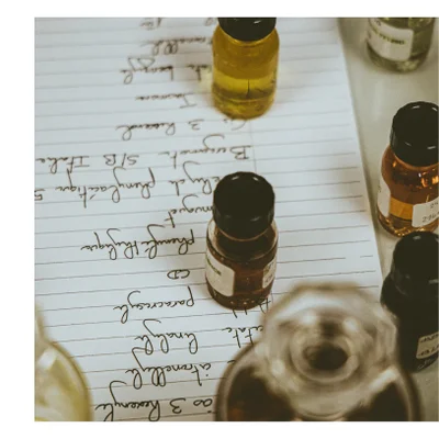 A close-up of several small bottles on a sheet of lined paper with handwritten text listing ingredients including Bergamote S/B Italie 5 Phenylacetique Muguet Phenyl CD Paracresol linalool citronellyl ao 3 Hoxenyl and text on bottle labels including ENCE 24 214.