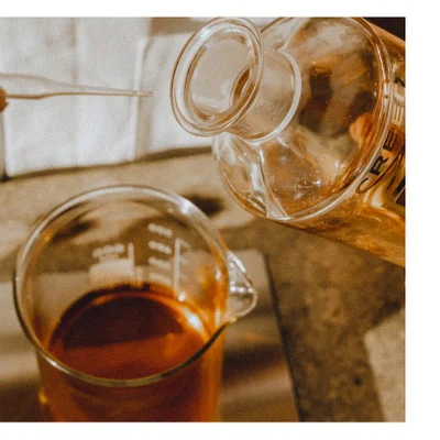 Close up of liquid being poured from a glass bottle labeled CREED into a glass beaker with a pipette in the background.