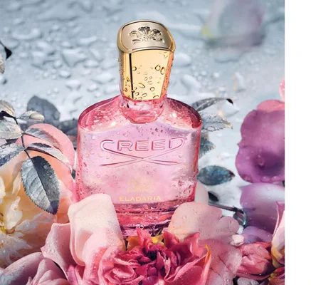 A pink Creed Eladaria perfume bottle with water droplets is surrounded by wet pink and purple roses and leaves.