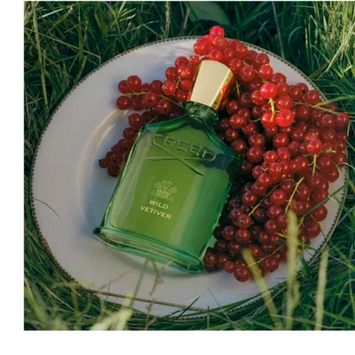 A green perfume bottle with a gold cap rests horizontally in lush green grass with some small white wildflowers and clover, displaying the brand name CREED and the scent name WILD VETIVER.