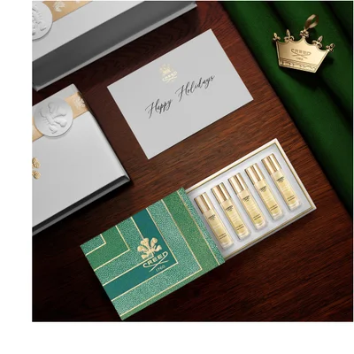 A Creed holiday gift set is displayed on a wooden surface, featuring an open green and gold box containing six miniature perfume bottles, a white closed gift box, a white greeting card, and a golden crown ornament resting on green fabric. The greeting card text is Happy Holidays and CREED 1760, the open gift box displays CREED 1760, the crown ornament also has CREED 1760, and the miniature perfume bottles are each labeled CREED 1760, 10 ML 0.33 FL.OZ, EAU DE PARFUM or EAU DE COLOGNE, MADE IN FRANCE, with individual fragrance names.
