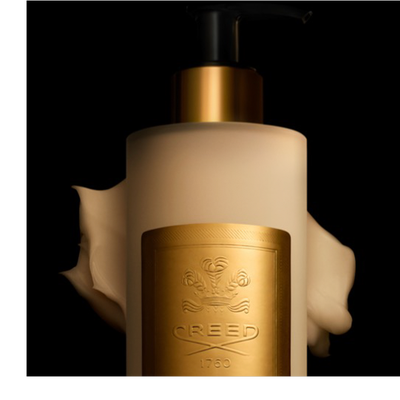 A creamy beige lotion bottle with a black and gold pump dispenser and a golden label featuring a crest and the text CREED 1760 has product swirled around it against a black background.