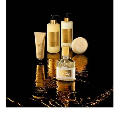 A set of gold-toned Creed Aventus for Her luxury products, including a perfume bottle with an equestrian emblem, two pump bottles, a hand cream tube, and a round soap bar, are arranged on a reflective black surface with golden ripples, showing text such as CREED 1760, AVENTUS FOR HER, HAND CREAM CREME MAINS, LIQUID SOAP SAVON LIQUIDE POUR LE CORPS, and BATH & BODY MILK LAIT DE BAIN ET CORPS.