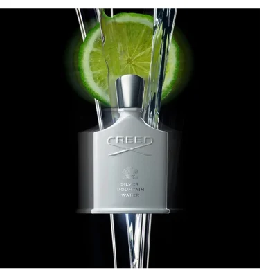 A black Creed perfume bottle with a dark cap rests on dark green fern leaves against a black background, with visible text Creed and Green Irish Tweed on the bottle.