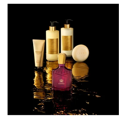 A red Creed Carmina perfume bottle with a gold cap, a beige hand cream tube, two beige pump bottles for hand and body wash and lotion, and a round beige soap bar are displayed on a dark reflective surface with golden liquid effects; visible text on the items includes Creed, 1760, Carmina, Hand Cream, Creme Mains, Hand & Body Wash, Lavant Liquide, Hand & Body Lotion, and Lait Riche.