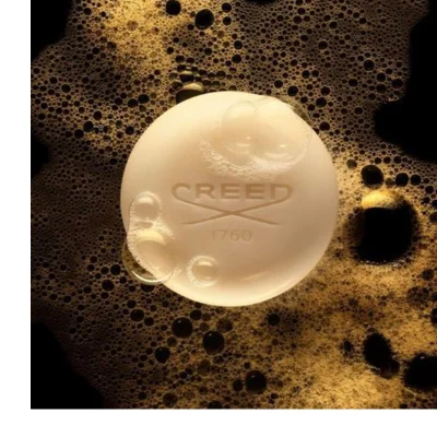A round, light-colored bar of soap with some bubbles on its surface rests in dark golden-brown foam, featuring the embossed text CREED with a logo and 1760, against a black background.