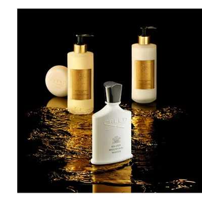A white Creed Silver Mountain Water perfume bottle, a light yellow pump bottle of hand and body wash, a cream-colored pump bottle of hand and body lotion, and a round bar of soap are artfully arranged on a shimmering gold liquid surface against a black background, displaying text that includes CREED, 1760, SILVER MOUNTAIN WATER, HAND & BODY WASH, and HAND & BODY LOTION.