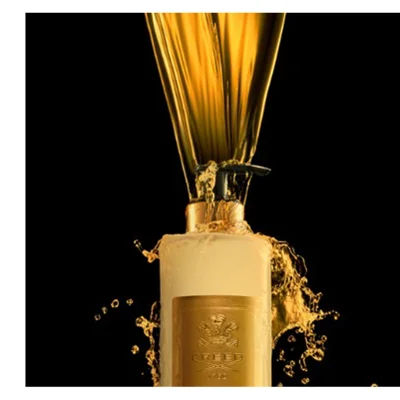 Golden liquid pours over a cream-colored bottle with a black pump, splashing around the top, revealing a golden label with a crest and the text CREED 1760.
