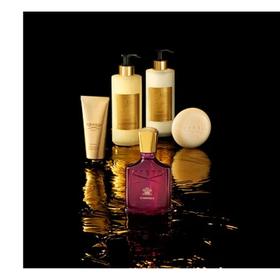 A luxurious collection of five Creed Carmina fragrance and body products is arranged on a glossy black surface with golden liquid reflections: a prominent dark red perfume bottle with a gold cap, a golden tube of hand cream, two tall golden pump bottles for hand and body wash and lotion, and a round cream-colored soap, bearing the visible text Creed 1760 Carmina, Creed 1760 Carmina Hand Cream, Creed 1760 Carmina Hand & Body Wash, Creed 1760 Carmina Hand & Body Lotion Lait de Corps, and Creed 1760.